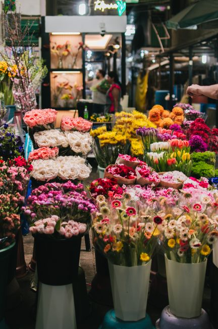 A vibrant floral market display featuring a variety of fresh flowers arranged in multiple styles. In the foreground, there are tall white vases filled with colorful daisies, tulips, and chrysanthemums