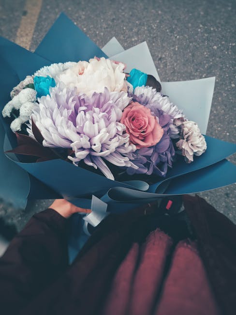A hand holds a beautifully arranged floral bouquet featuring a mix of fresh flowers including large white and lavender chrysanthemums, soft pink roses, and smaller blue accent flowers. The bouquet is
