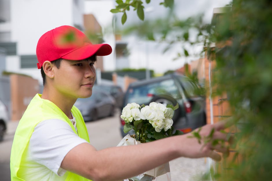 A delivery person wearing a red cap and neon yellow vest is placing a bouquet of white hydrangeas wrapped in white paper into a flower holder next to a green leafy plant outside on a street. The backg