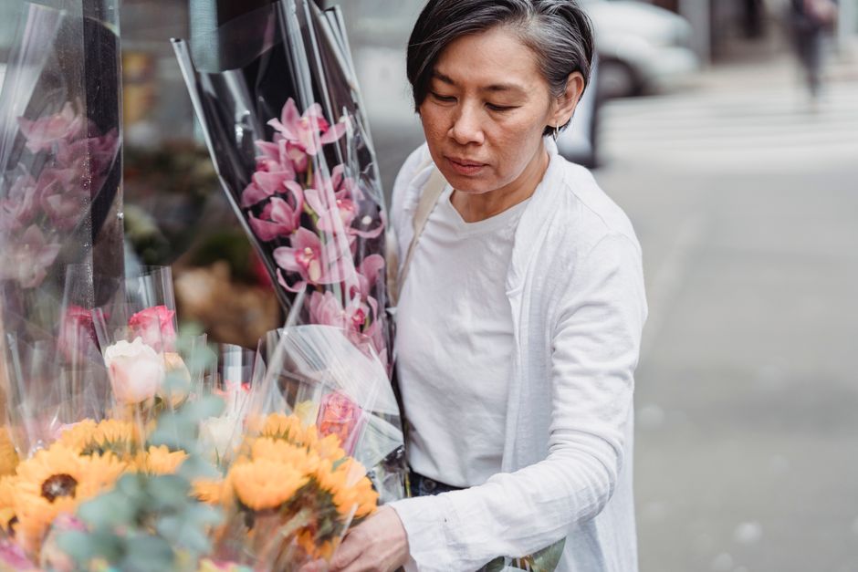 A woman with short, dark hair and a white shirt is examining a collection of fresh flower bouquets outside a florist shop. The arrangements feature pink orchids in clear plastic wrapping, along with y