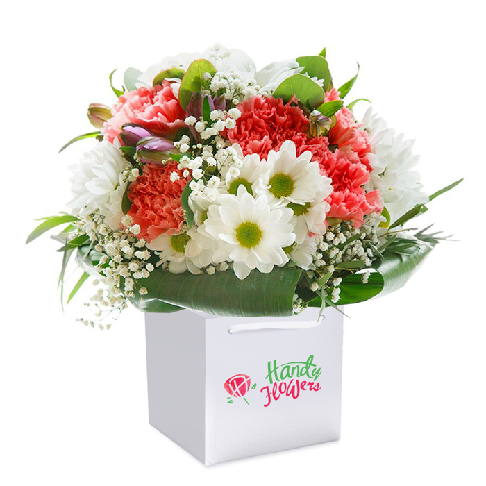 In this thoughtfully composed bouquet, flourishing within a square white gift bag, every flower and leaf channels the contemporary yet welcoming spirit of West Wickham. Ruffled petals of coral and soft peach carnations cluster at the bouquet's center, offering texture and a gentle, alluring scent reminiscent of well-kept private gardens around the Langley Park area. Rich red carnations punctuate the softer tones with bursts of vibrancy, while snow-white daisies-each with a glistening lime-green heart-bring brightness and balance. Throughout the arrangement, clouds of delicate baby's breath shimmer, lending an ethereal lightness. Along one edge, subtle purple-tipped buds whisper of blooms yet to come, creating a sense of anticipation and depth. Surrounding all of this, deep green, broad foliage sweeps around the flowers with confidence, both supporting the blooms and forming an elegant visual collar, much like the mature trees that line the quieter roads off Wickham Court Road. The bouquet's rounded, abundant profile is a statement of care and local pride, expertly arranged and ready to be presented for occasions ranging from birthdays to anniversaries-or simply to extend a thoughtful gesture to someone special in BR4. The white bag, with strong, looped ribbon handles, sits set against a seamless white backdrop, drawing all attention to the artistry within. It is a floral vignette that captures the essence of beauty, quality, and the personal touch found at Flowers West Wickham.
